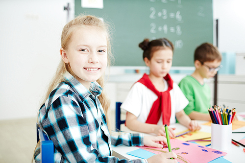 Happy blond schoolgirl smiling at you while sitting by desk and drawing at lesson