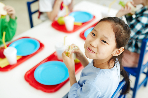 Pretty Asian girl sitting at table with classmates in primary school canteen, eating sandwich and smiling at camera cheerfully