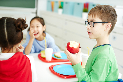 Caucasian boy in glasses eating apple while having lunch with classmates in elementary school cafeteria