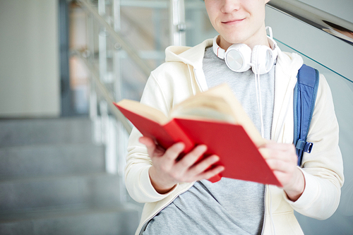 Young casual student with headphones on neck reading book in red cover by staircase