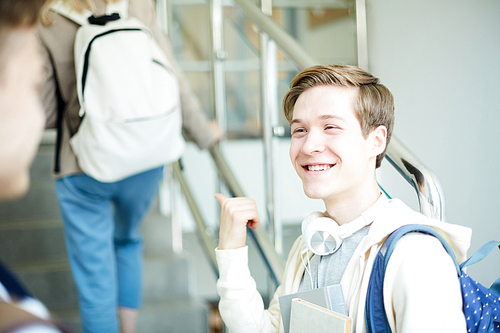 Happy guy pointing at girl with backpack moving upstairs while discussing her with friend