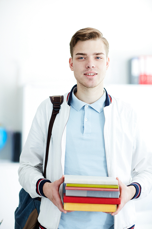 Handsome student with stack of books looking at you in college library