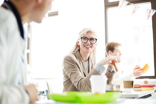 Joyful blond girl with sandwich looking at one of groupmates at lunch break in classroom