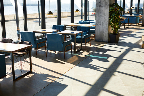 Wooden tables and blue chairs standing along windows in cafe by riverside on sunny day