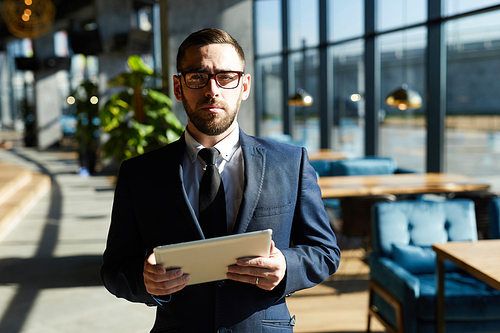 Bearded businessman in suit and eyeglasses standing in cafe while using touchpad