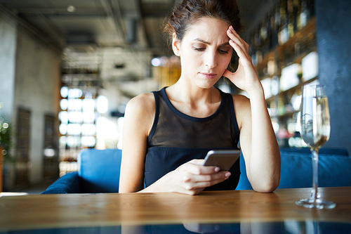 Worried woman touching her head while sitting by table in cafe, messaging in smartphone and waiting for someone