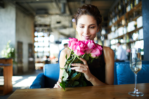 Happy young woman looking at bunch of pink roses in her hands while sitting by table in cafe