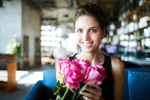 Cheerful pretty girl with bunch or roses looking at you while relaxing in cafe or restaurant