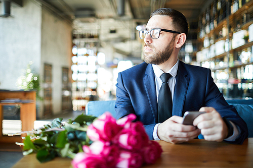 Young serious man with smartphone waiting for date in restaurant with bunch of roses near by