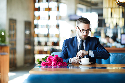 Elegant man having coffee while sitting by table in restaurant, looking at watch and waiting for girlfriend
