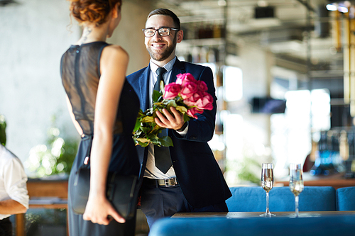 Happy young man giving bunch of pink roses to his girlfriend during romantic date in restaurant