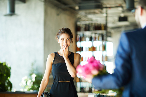 Amazed young woman touching her mouth while looking at bunch of flowers in her boyfriend hands