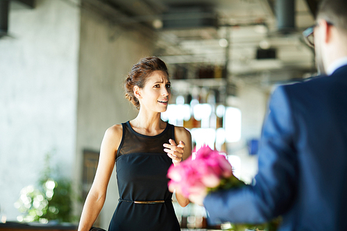 Surprised young woman in black dress pointing at bunch of roses held by her boyfriend