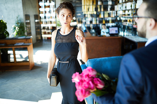 Annoyed girl in elegant dress talking to man with bouquet of pink roses in restaurant