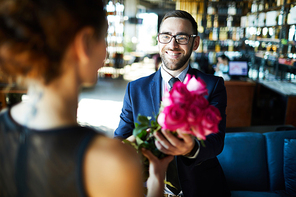 Happy man in suit and eyeglasses giving rose bouquet to his girlfriend in restaurant