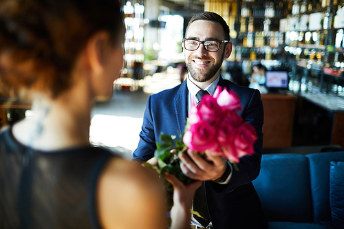 Happy man in suit and eyeglasses giving rose bouquet to his girlfriend in restaurant