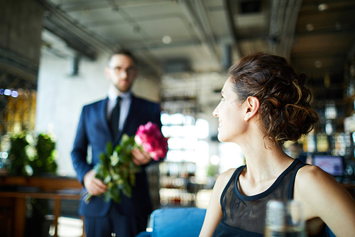 Young brunette sitting in restaurant and looking at her boyfriend carrying rose bouquet