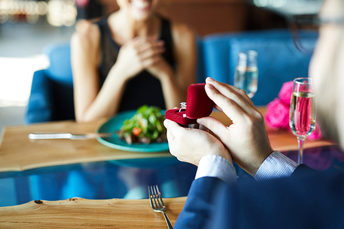 Diamond ring in small red velvet box held by young man making proposal to his girlfriend in restaurant