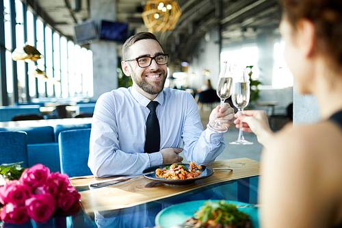 Young cheerful man clinking his flute with that of his girlfriend over served table on romantic date in restaurant
