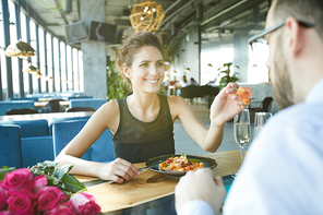 Cheerful young woman giving her food to boyfriend to taste during romantic dinner in restaurant