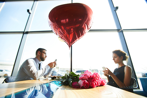 Couple of valentines sitting by served table with bunch of roses and red heart-shaped balloon and using their gadgets