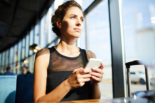 Pretty businesswoman with smartphone looking through window on sunny day while sitting in airport lounge