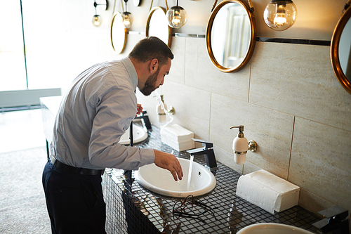 businessman in white shirt and pants leaning over sink in lavatory while refreshing mouth