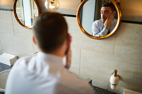 Reflection in mirror of young businessman taking care of his face in toilet or bathroom