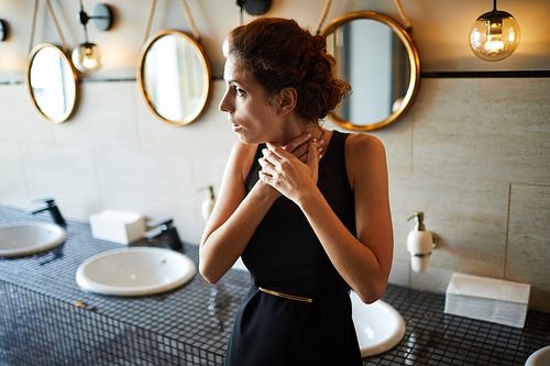 Young brunette woman in elegant black dress touching her throat while standing in toilet