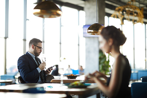 Young contemporary businessman with flute of champagne texting in smartphone while sitting by table in restaurant