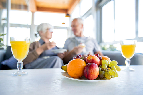 Blurred portrait of modern senior couple enjoying breakfast in cafe, focus on fruit bowl and two glasses of orange juice on table in foreground