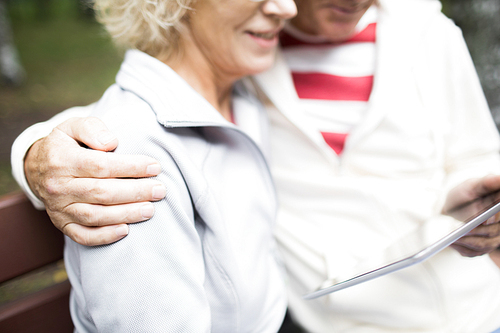 Mature man with touchpad embracing his wife while both sitting in park and watching online video