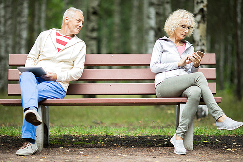 Senior man with touchpad looking at female texting in smartphone on other side of bench