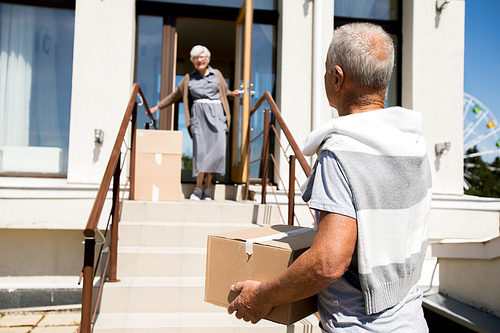 Portrait of modern senior couple unloading cardboard boxes while moving to new house, focus on man carrying box