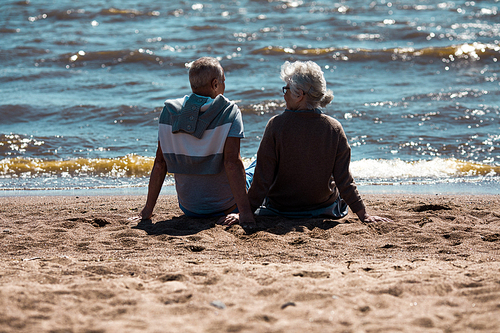 Back view of senior couple sitting on sandy beach, talking and relaxing in front of waterside