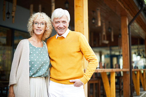 Smiling fashionable senior couple standing on veranda and , handsome man embracing girlfriend in glasses