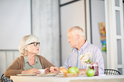 Modern senior couple sitting by table in modern cafe, having rest and talking by cup of tea