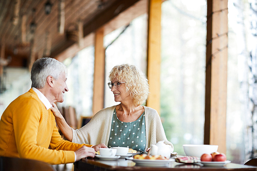 Happy beautiful senior wife with curly hair wearing glasses touching shoulder of husband and supporting him while they talking at breakfast on porch