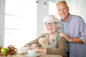 Happy aged woman with bunch of flowers and her husband near by  while sitting by table