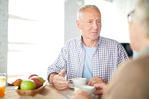 Senior man in casualwear sitting by table, having tea and talking to his wife at breakfast