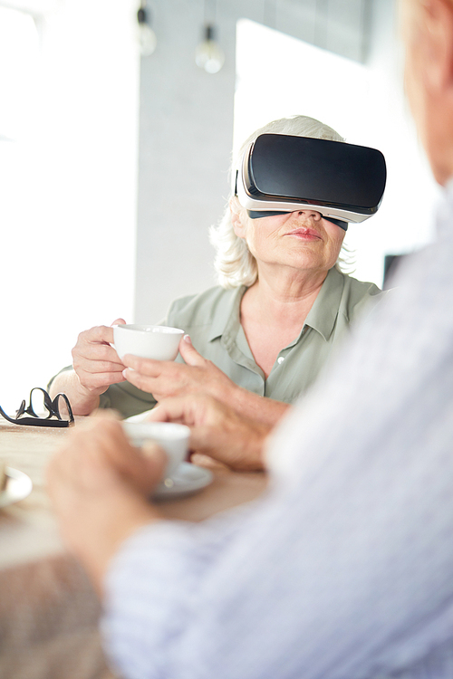 Senior woman with cup of tea wearing vr headset while having talk to her husband