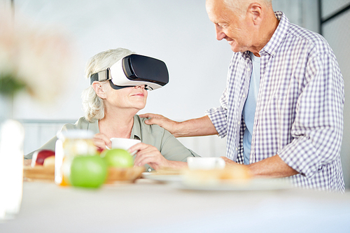 Senior man standing by his wife wearing virtual reality goggles during conversation in the kitchen