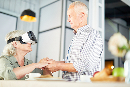 Aged female with vr headset and her husband holding by hands during breakfast