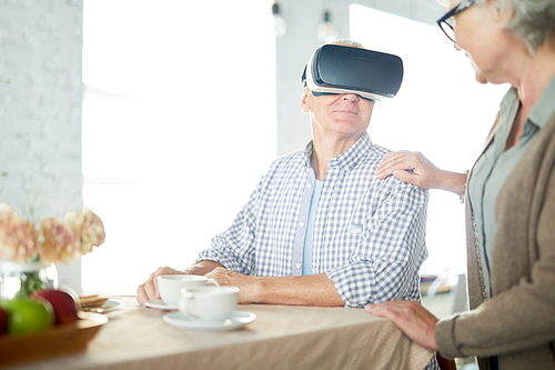 Aged man in vr goggles sitting by table, having tea or coffee and talking to his wife by breakfast