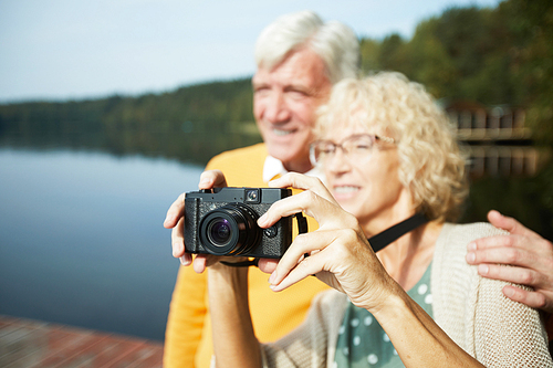 Positive beautiful senior couple being on vacation standing on pier and photographing nature on camera, handsome elderly man embracing beloved woman