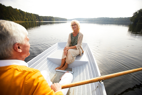 Smiling senior lady in glasses looking with love and tenderness at boyfriend who rowing boat and enjoying date on boat