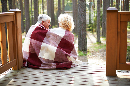 Rear view of happy senior couple covered with checkered blanket talking and sitting on porch of country house