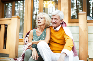 Positive dreamy senior lovers in casual clothing under plaid sitting on porch and looking into distance, happy man embracing his lady at veranda