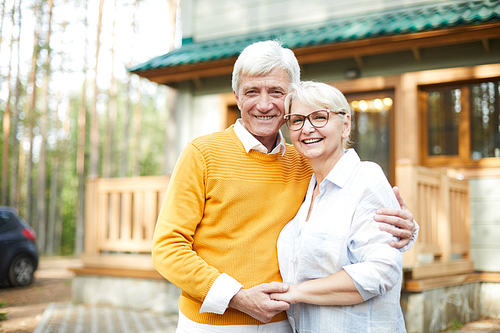 Forest house of grandparents: cheerful excited senior couple embracing each other and laughing while  and standing against cottage