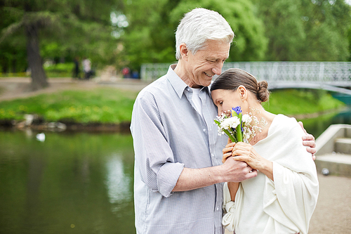 Amorous seniors holding bunch of wildflowers while woman leaning on her husband shoulder in park